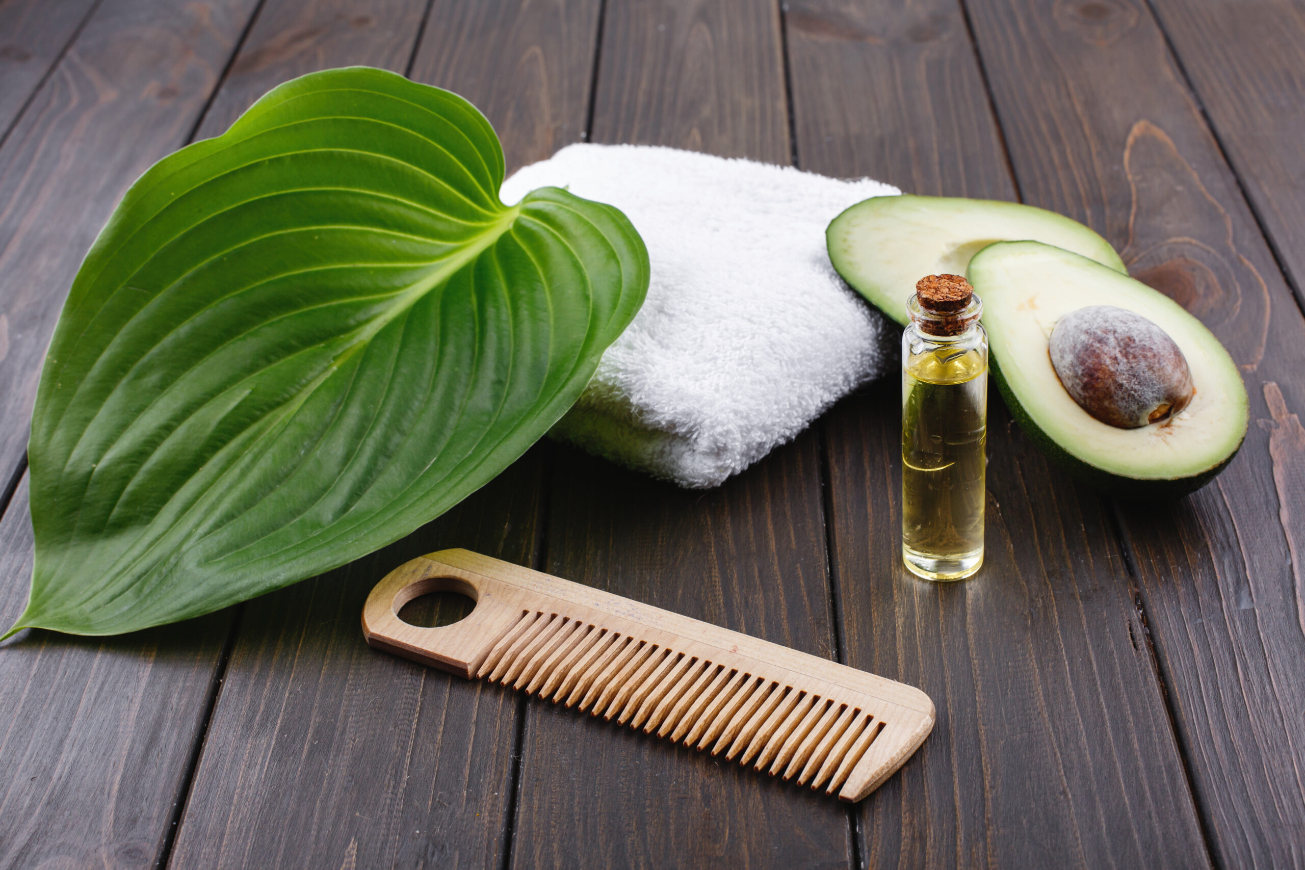 aceites esenciales para el cabello. una mesa con una toalla blanca, un peine una hoja , un aguacate y un bote de aceite esencial para el cabello